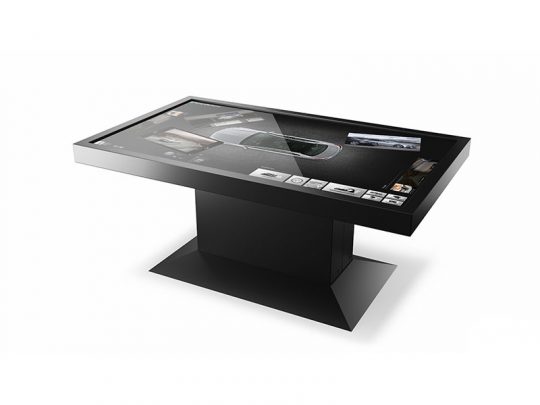 86 Zoll 4K/UHD Touch Table – Screensource AVRG86T shown with a digital car customization interface on its large, horizontal touchscreen; features a sleek, modern black pedestal base. The display presents a top-down view of a car and menu options for interactive customization.