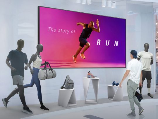 A man in sportswear views a retail display featuring athletic mannequins, gym bags, running shoes, and a large 110-inch LG 110UM5J-B digital signage display for rent. The screen shows an image of a runner with the text “The story of RUN.” This setup highlights sports apparel and equipment in a modern store environment.