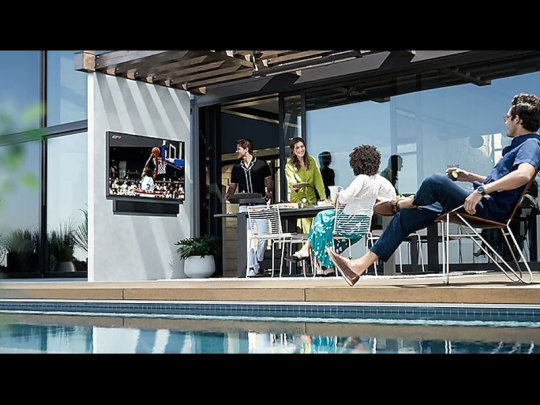 Four people relax by a poolside patio, enjoying a basketball game on a 75 Zoll Outdoor 4K QLED-TV – Samsung The Terrace (rented); some are seated at a table and one lounges with a drink, creating a bright, casual atmosphere ideal for outdoor TV entertainment.
