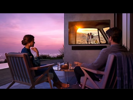 A woman and man watch a beach scene at sunset on the 75 Zoll Samsung The Terrace Outdoor 4K QLED-TV, with city lights in the background and drinks on the table between them.