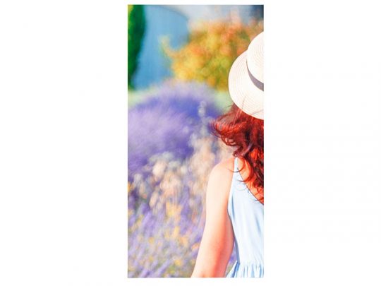 Woman with long red hair wearing a light blue summer dress and straw hat, standing outdoors with blurred purple and green foliage in the background. Perfect for promoting casual spring or summer fashion.