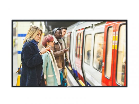 A group of commuters in winter clothing stand on a subway platform, looking at their phones, as a train with red and white doors arrives indoors. Subway station scene showing urban daily commute.