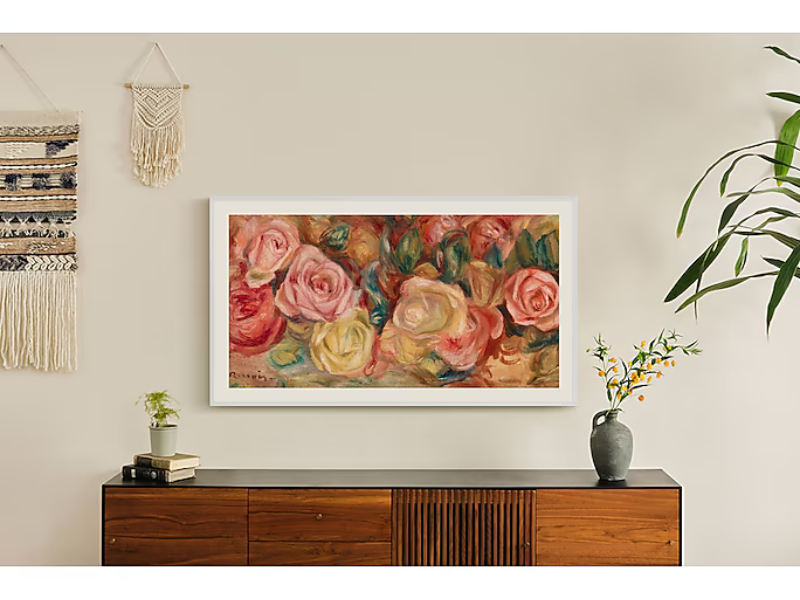 Samsung The Frame 43-inch 4K UHD QLED TV mounted on a beige wall above a wooden sideboard decorated with books, a small plant, a flower vase, and a woven wall hanging; a large green plant stands beside the cabinet.