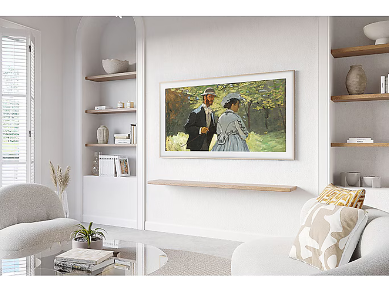 A modern living room with neutral decor features the Samsung The Frame 43 Zoll 4K UHD QLED-TV mounted on a white wall above a floating wooden shelf, framed by built-in shelves and comfortable seating. The TV blends seamlessly with the stylish surroundings, combining art and entertainment.