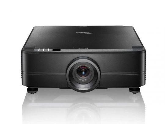 Black home theater projector with a central lens, positioned on four small feet, displayed on a white reflective surface against a plain white background.