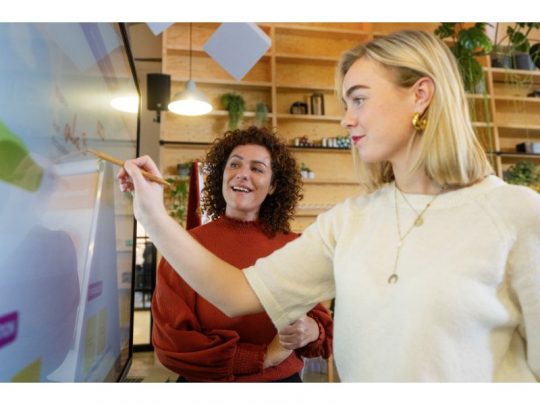 Zwei Frauen arbeiten gemeinsam an einem Legamaster EXPLORE 2 EXP-6510 65 Zoll Touchdisplay in einem modernen Büro. Eine Frau bedient das interaktive Whiteboard mit einem Stift, die andere lächelt, im Hintergrund sind Regale und Pflanzen zu sehen. Ideal für innovative Teamarbeit und digitale Meetings - jetzt Legamaster EXPLORE 2 EXP-6510 (Neuware) kaufen.