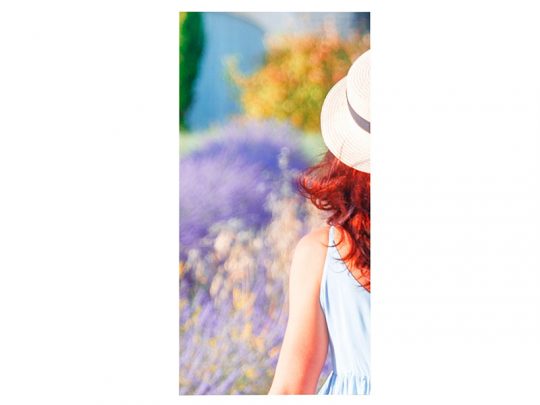 Eine Person mit langen rötlichen Haaren trägt ein hellblaues Kleid und einen Strohhut. Sie steht im Freien und blickt von der Kamera weg; im Hintergrund sind unscharfe Lilien und grüne Pflanzen zu sehen. Ideal für Sommermode oder Outdoor-Lifestyle-Kampagnen.